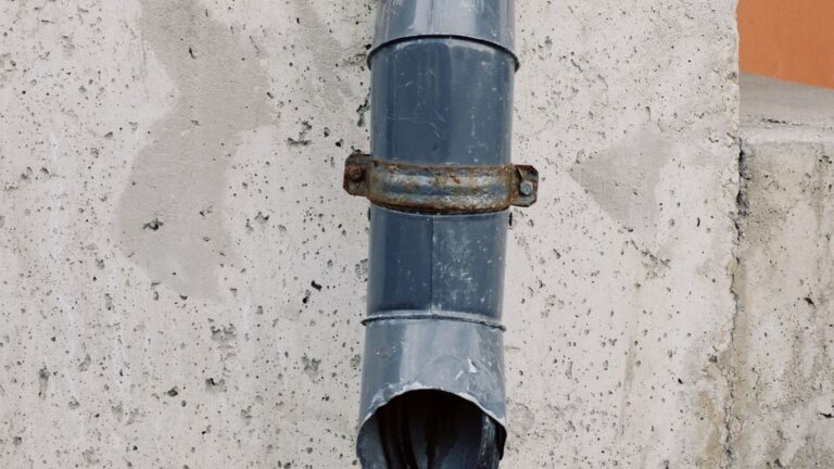 How Far Should Downspouts Drain From the House? Downspout (drain pipe) running down a house's concrete exterior wall.