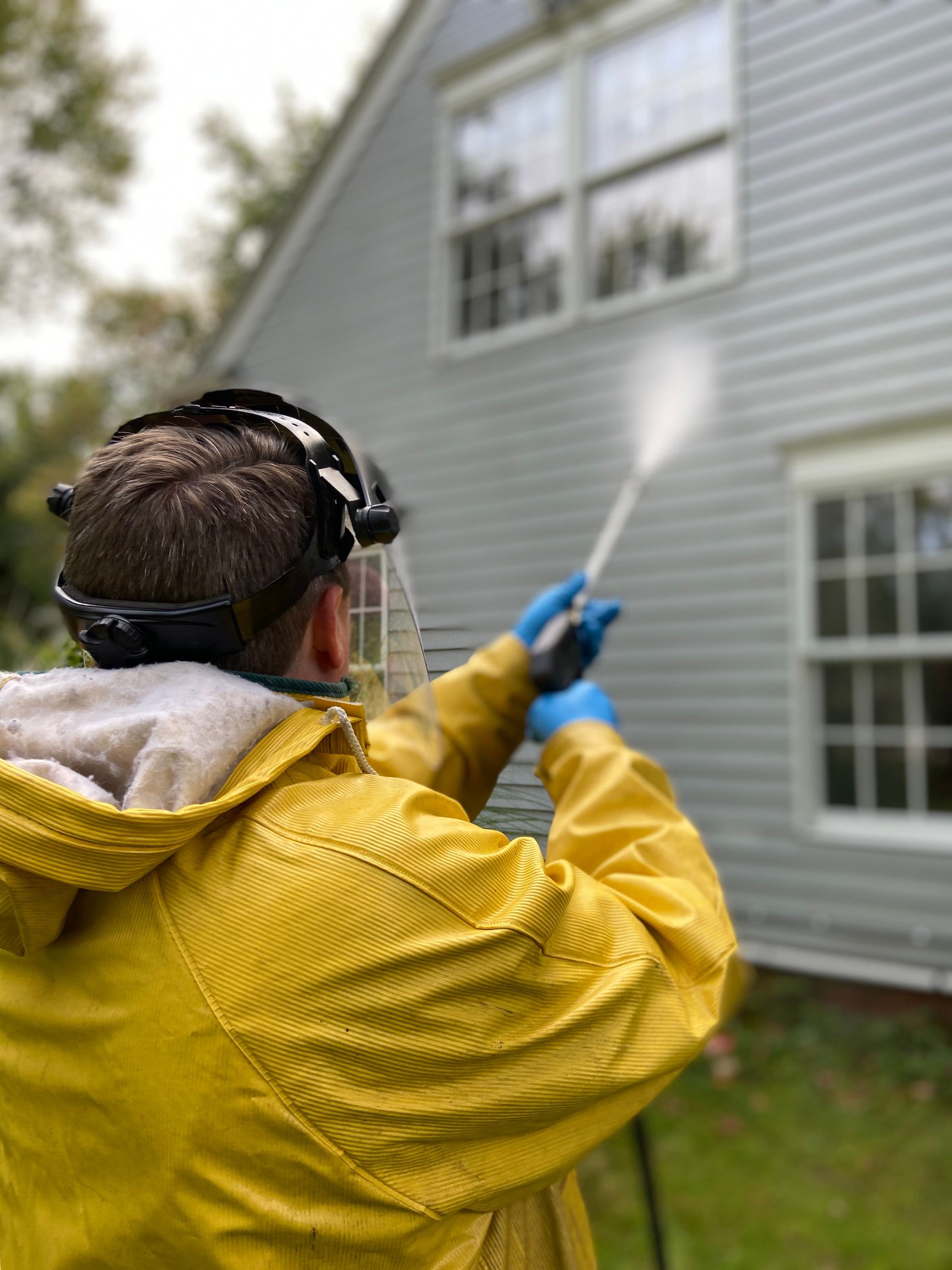 A worker in a yellow raincoat, face shield, and gloves rinsing house siding with a pressure washer