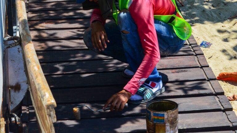 Worker crouching on wooden deck boards applying stain with a small roller, an open paint can beside him