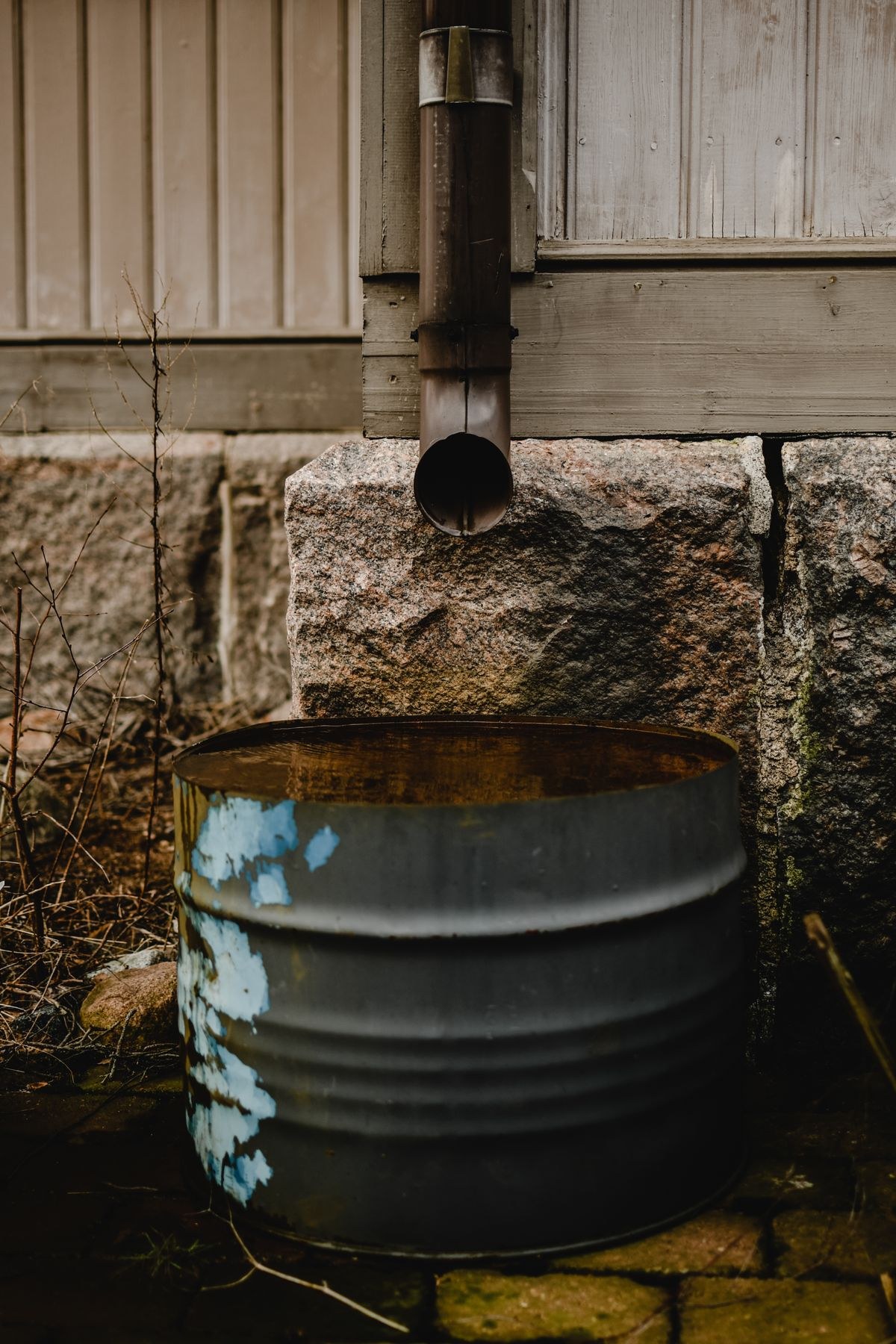 A downspout emptying into a rain barrel beside a stone foundation wall