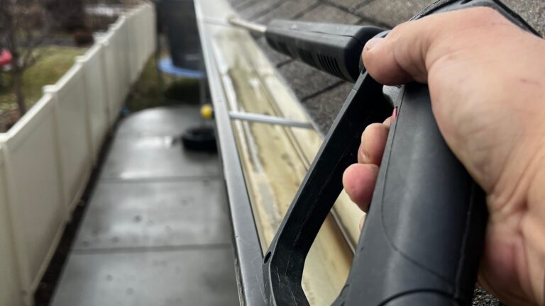 Questions to Ask a Gutter Cleaning Company Before Hiring Close-up of a worker pressure-washing a house gutter from the roof during a professional gutter cleaning visit