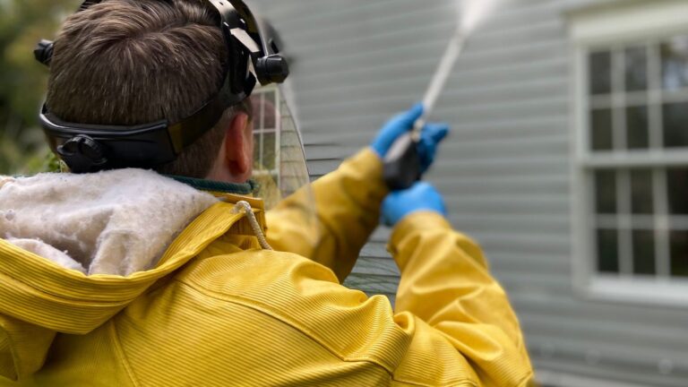 Soft Washing Cost Factors: What Affects a House-Washing Quote? Professional in a yellow raincoat washing house siding with a low-pressure sprayer