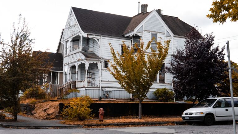 A weathered white wooden house with autumn trees and fallen leaves on a suburban street in fall
